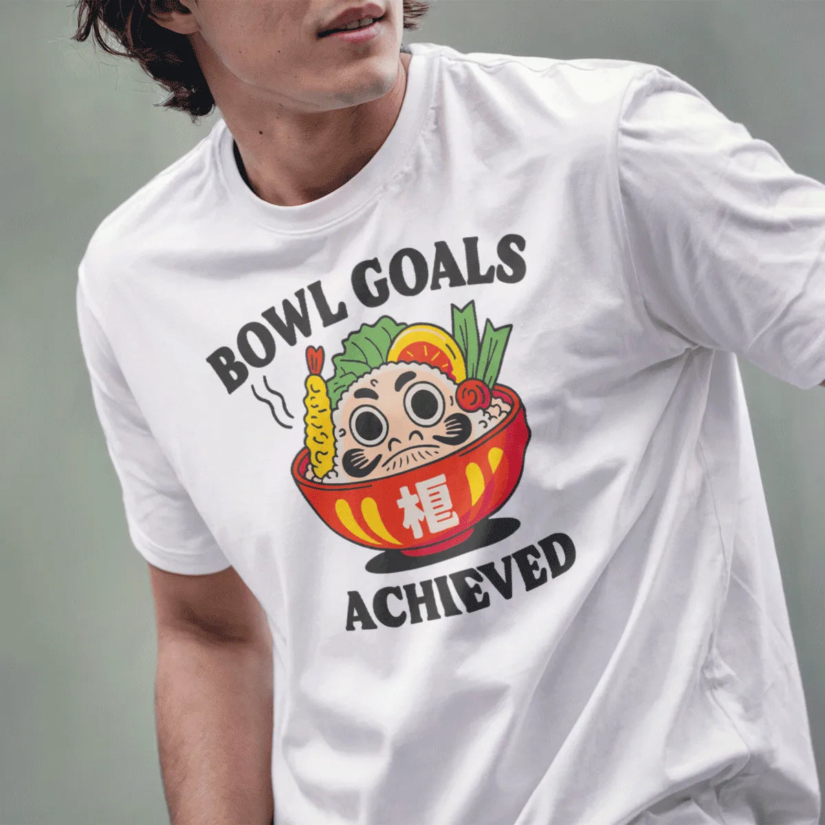 Male model in white t-shirt with Daruma bowl, sushi, and “BOWL GOALS ACHIEVED” text, positive messaging.